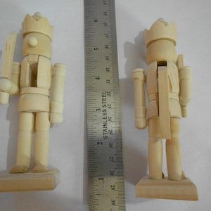 May include: Two unfinished wooden nutcracker figurines, each with a crown, holding a sword or a staff. The figurines are approximately 13 cm tall and stand on rectangular bases. A stainless steel ruler is placed between them.