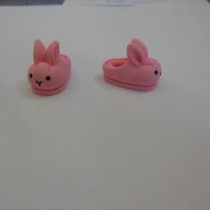 May include: A pair of pink bunny-shaped slippers. The slippers have black eyes and a small black nose.