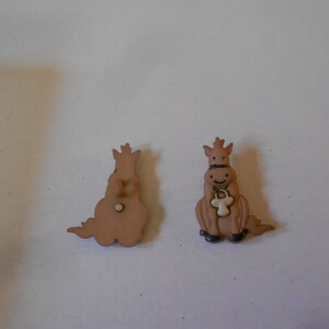 May include: Two brown plastic buttons shaped like horses. One button is a side view of a horse with a white halter and the other button is a back view of a horse.