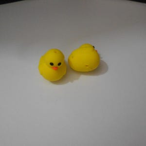 May include: Two small, yellow chick figurines with black eyes and orange beaks. The chicks are sitting on a white surface. The figurines are likely Easter or spring-themed decorations.