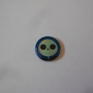 Può includere: Un bottone blu e verde con un'immagine di cartone animato di Jack Skellington dal film The Nightmare Before Christmas.