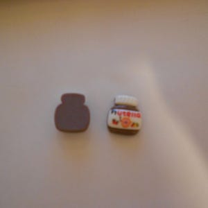 Peut inclure: Deux breloques miniatures en forme de pot marron. L'une est une forme marron pleine, l'autre est un petit pot de Nutella avec un couvercle blanc et une étiquette avec le logo Nutella et des accents floraux.