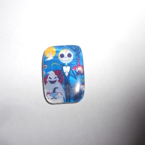Può includere: Un cabochon di vetro blu e bianco con un'immagine colorata di Jack Skellington del film L'incubo prima di Natale. L'immagine include anche Sally e Zero.