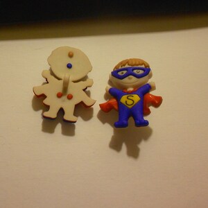 May include: Two plastic buttons featuring cartoon characters. One button is a white figure with red and blue accents. The other button is a blue superhero figure with a red cape and a yellow 'S' on the chest.