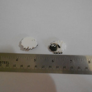 May include: Two small, white sheep figurines with black and green details. The sheep are positioned on a white surface next to a stainless steel ruler. The ruler shows measurements in inches.