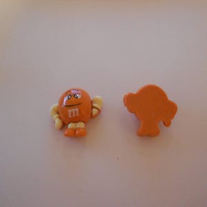 以下が含まれることがあります： オレンジ色のM&M'sキャンディー2つ。1つは「m」のプリント入り、もう1つは木の形。キャンディーのキャラクターは白い手袋、靴、白いアウトライン。キャンディーは白い表面にあります。