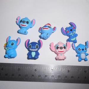 以下が含まれることがあります： 様々なポーズの、小さくてカラフルなスティッチのフィギュアが8体。フィギュアは主に青色で、耳と鼻にピンク色のアクセントがあります。サンタ帽をかぶったフィギュアも。フィギュアの下には定規が置かれています。