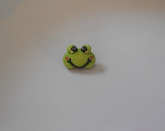 Könnte beinhalten: Ein grünes Froschgesicht mit schwarzen Augen, einem schwarzen Lächeln und rosa Wangen. Das Froschgesicht ist ein Knopf aus Plastik.