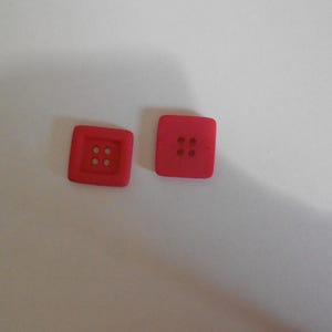 May include: Two square, bright red buttons with four holes each. The buttons are made of a solid material and are set against a white background. These buttons are suitable for sewing projects and crafts.