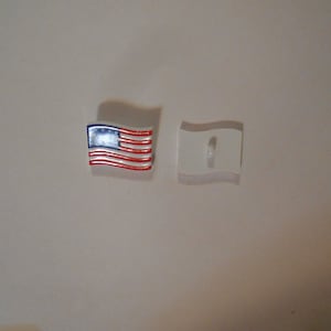 Puede incluir: Un botón de plástico blanco con un diseño de bandera estadounidense. El botón tiene un agujero en el centro para coser.