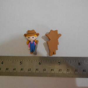 Può includere: Due piccole statuette di contadina. Una è una figura dettagliata e colorata di una ragazza con un cappello marrone, una tuta blu e un rastrello. L'altra è una silhouette marrone solida della stessa figura. Un righello di metallo è in primo piano.