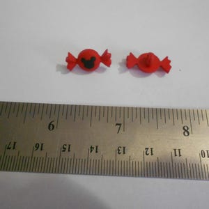May include: Two red, candy-shaped embellishments with a black Mickey Mouse silhouette on one. A metal ruler with centimetre markings is in the foreground. The embellishments are approximately 2 cm in length.