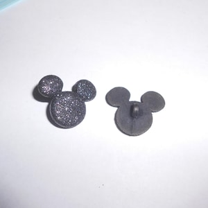 Puede incluir: Dos botones grises brillantes con forma de Mickey Mouse. Un botón muestra el frente con purpurina, el otro muestra la parte posterior con un ojal. Los botones están sobre una superficie blanca.