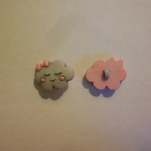 Può includere: Due bottoni di plastica rosa e grigi a forma di nuvole. Una nuvola ha un fiocco rosa e un viso sorridente con gli occhi chiusi. L'altra nuvola ha un foro al centro per cucire.