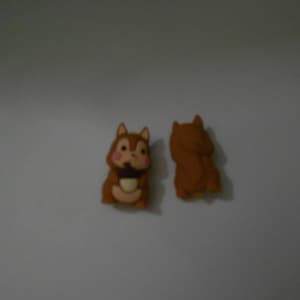 May include: Two brown squirrel figurines. One squirrel is facing forward, holding a nut, with pink cheeks. The other squirrel is facing away. Both have pointed ears and fluffy tails. The background is a plain, light gray.