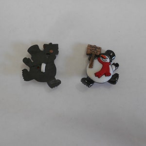 May include: Two plastic Christmas buttons. One is a black snowman with a top hat and a sign that says "Snow Sale". The other is a black bear wearing a top hat.