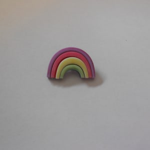 Op de afbeelding: Een kleine regenboogkleurige plastic pin met vijf kleurbanden: paars, roze, geel, groen en blauw.