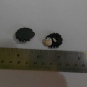 May include: Two small, black sheep-shaped embellishments with white and tan details. One is a complete sheep, the other is a dark gray, cloud-like shape. A metal ruler is in the foreground.