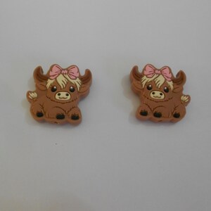 以下が含まれることがあります： ピンクのリボンが付いた茶色のハイランド牛のチャームが2つ。チャームはゴム製で、漫画のような外観です。