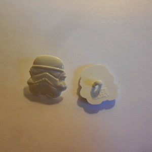 Puede incluir: Botón de plástico blanco con una imagen en relieve de un casco de Stormtrooper de Star Wars. La parte posterior del botón tiene el texto "OFL" impreso.