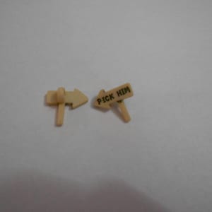 Puede incluir: Dos pequeñas señales direccionales beige. Una señal tiene una flecha que apunta hacia la derecha. La otra señal dice "PICK HIM". Las señales están hechas de un material de color claro y están sobre un fondo liso.