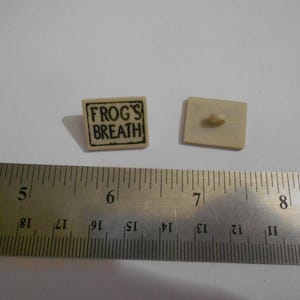 May include: Two small, off-white square buttons. One button has the text "FROG'S BREATH" in a black, rectangular frame. The other button has a small, rounded protrusion on the back. A metal ruler with inch markings is in the foreground.