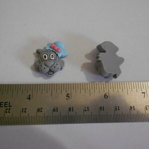 May include: Two gray cat-shaped buttons. One button features a cat face wearing a blue hat with a pink bow. The other button is a solid gray cat shape. A metal ruler with inch measurements is in the foreground.