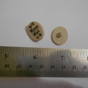 May include: Two small, off-white buttons are displayed next to a metal ruler. One button is oval-shaped and has the words "Worm's Wort" printed on it in black script. The other button is round with a shank on the back. The ruler shows measurements in inches.