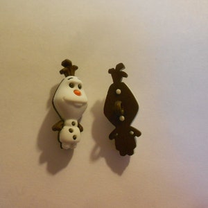 May include: Two decorative buttons shaped like Olaf from Frozen. One button is a full-color Olaf, the other is a brown silhouette. The buttons have a white body, brown arms, and a carrot nose.