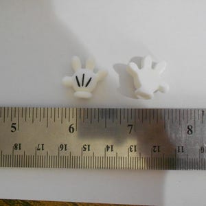 May include: Two small, white, cartoon-style hand-shaped embellishments with black lines on the fingers. A metal ruler with inch markings is in the foreground, with the hands positioned near the 6-inch mark.