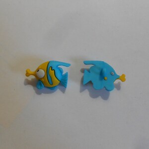 May include: Two blue and yellow plastic fish shaped buttons. One fish is facing left and the other is facing right.