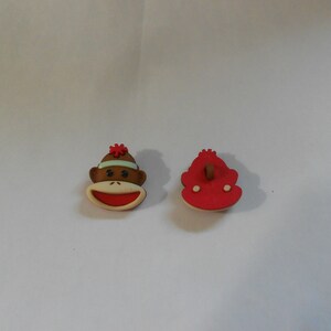 May include: Two plastic buttons, one with a monkey face and the other a solid red shape. The monkey button has a brown face, red mouth, and a green headband with a red flower on top.