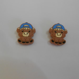 Op de afbeelding: Twee bruine cartoon-koe-charms met blauwe baseballpetten. De charms zijn gemaakt van rubber en hebben een 3D-ontwerp.