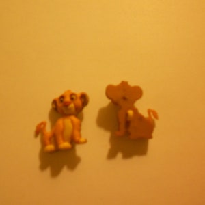 Puede incluir: Dos pequeñas figuras de plástico de Simba de la película El Rey León. La figura de la izquierda está de pie con la boca abierta y la cola enroscada detrás de ella. La figura de la derecha está sentada con la cola enroscada alrededor de su cuerpo.