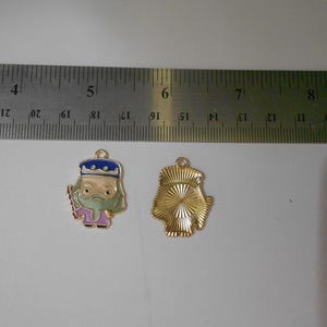 May include: Two metal charms. One is a colorful enamel pendant of a wizard with a blue hat, green beard, and purple robes. The other is a gold-colored charm with a textured back. A ruler is in the background.
