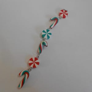 Puede incluir: Una pulsera festiva con diseños de bastones de caramelo y mentas. Los bastones de caramelo son rojos, blancos y verdes, mientras que las mentas son rojas y blancas. La pulsera está conectada por pequeños eslabones plateados, ideal para Navidad.