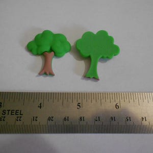 May include: Two small, green tree-shaped decorations with brown trunks. A stainless steel ruler with inch measurements is placed below the trees. The trees are likely for crafts or cake decorating.