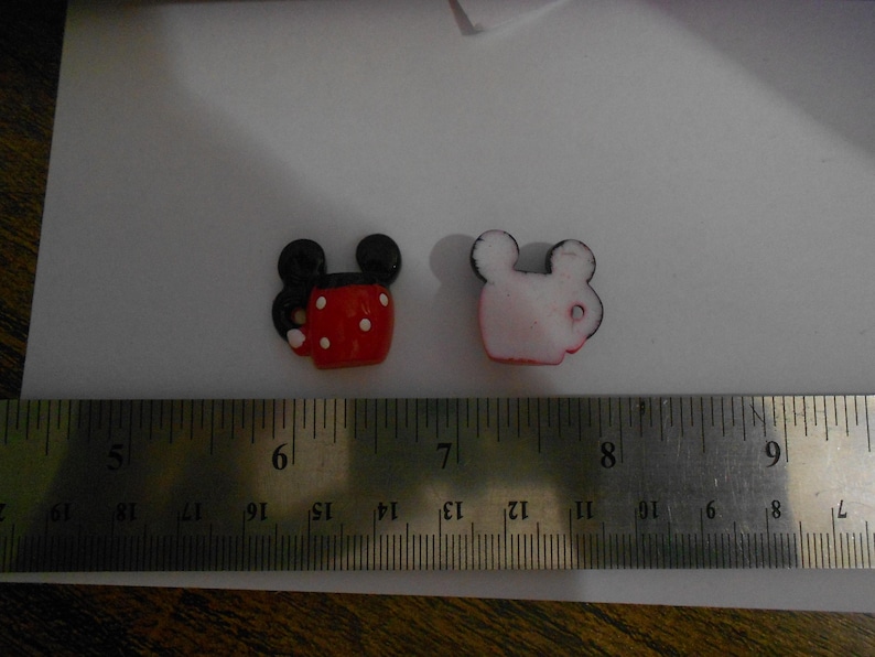 K&ouml;nnte beinhalten: Zwei kleine Mickey-Mouse-f&ouml;rmige Charms. Einer ist rot mit wei&szlig;en Punkten und schwarzen Ohren, der andere ist wei&szlig; mit schwarzem Rand. Die Charms liegen neben einem Lineal, das in Zentimetern misst.