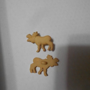 Puede incluir: Dos botones de plástico marrón en forma de alce. Los alces están de pie con la cabeza girada hacia un lado. Los botones tienen un diámetro de aproximadamente 2,5 cm.