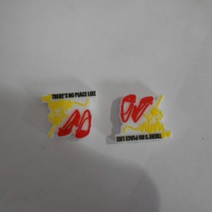 May include: Two shoe charms with the text "THERE'S NO PLACE LIKE" in black. One charm has red shoes and yellow accents, the other has red shoes and yellow accents. The charms are on a white background.