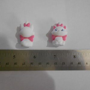 Puede incluir: Dos figuras de gato blanco con lazos y detalles rosas. Las figuras miden aproximadamente 2,5 cm de alto. Un gato tiene ojos azules. Las figuras están sobre una superficie blanca junto a una regla.
