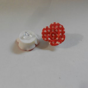 Puede incluir: Dos objetos decorativos: un objeto blanco en forma de flor con una gema transparente y una rosa roja con lunares blancos. La rosa tiene detalles de purpurina. Ambos objetos están sobre una superficie blanca.