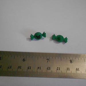 以下が含まれることがあります： 包まれたデザインと黒いハートが付いた2つの小さな緑色のキャンディー。センチメートル単位の目盛りが付いた金属製の定規が前景にあります。キャンディーの長さは約2cmです。