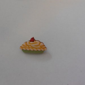 May include: A small, green pie crust filled with yellow and white frosting and topped with a red cherry.