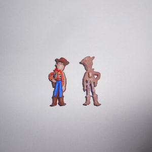 Peut inclure: Deux figurines en plastique de Woody de Toy Story. La figurine de gauche est en couleur, portant une chemise rouge et jaune, un jean bleu, un chapeau marron et des bottes marron. La figurine de droite est marron et montre l'arrière de la figurine.