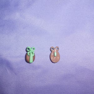 Op de afbeelding: Twee kleine plastic knopen, een bruin en een groen met een strik. De knopen zijn eivormig.