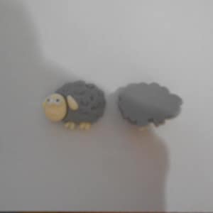 May include: Two gray sheep-shaped decorative embellishments. One is a complete sheep with a gray body, a yellow face, and yellow feet. The other is a gray sheep body without the head or feet. These are likely craft supplies.