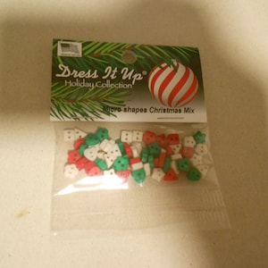 Puede incluir: Una bolsa de plástico transparente llena de microdecoraciones navideñas. La etiqueta dice "Dress It Up Holiday Collection" con un adorno a rayas rojas y blancas. Las decoraciones son rojas, verdes y blancas, en formas como mitones, árboles y muñecos de nieve.