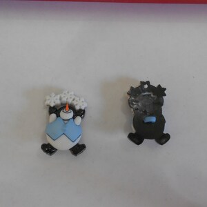 Op de afbeelding: Twee knoppen met een sneeuwman-thema. De ene knop heeft een sneeuwman met een blauwe vest en een witte hoed met sneeuwvlokken. De andere knop is zwart en heeft een sneeuwman-silhouet.