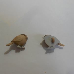 May include: Two brown plastic bird buttons with a white back. The bird on the left is facing forward and the bird on the right is facing the opposite direction.